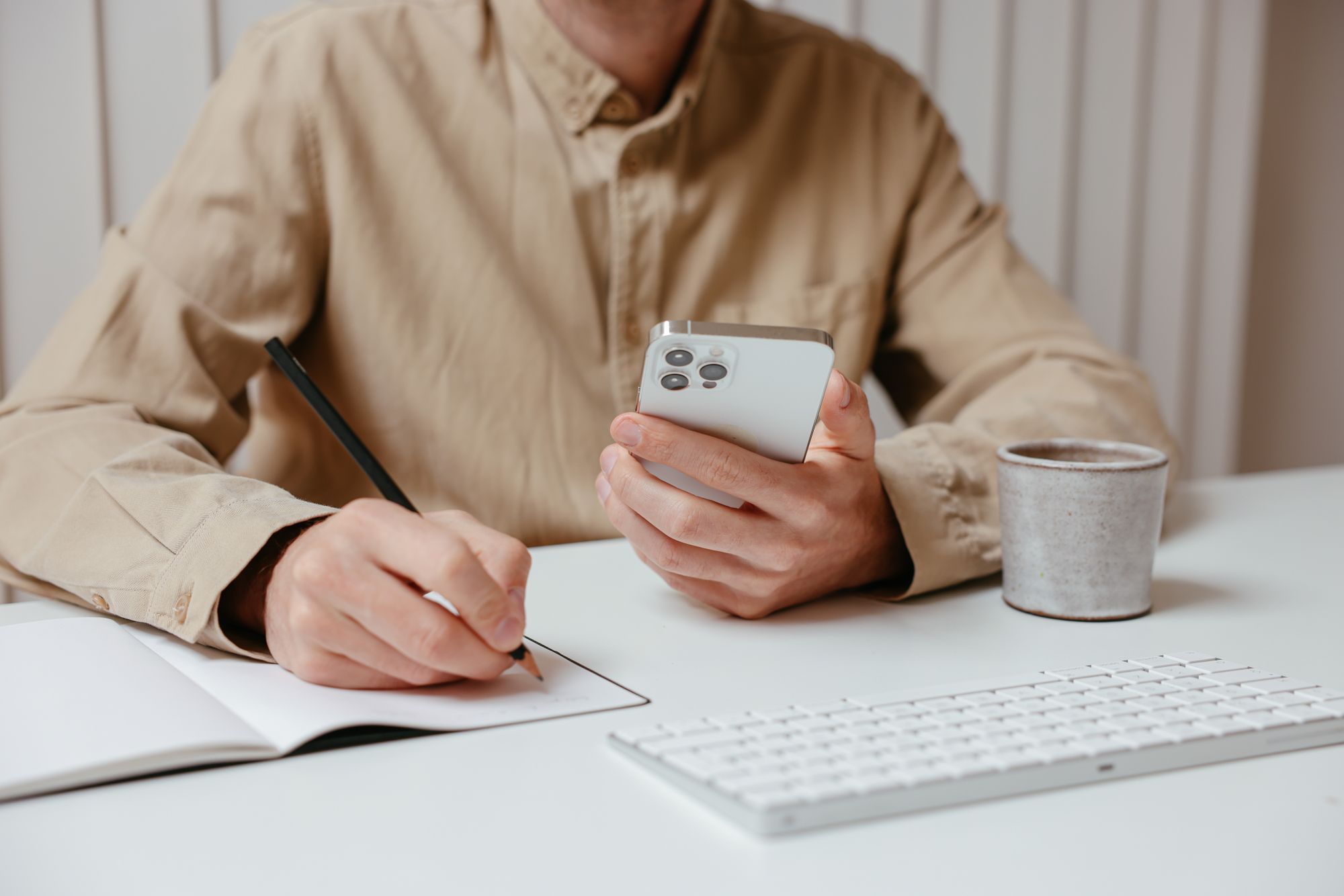 Ein Mann hält am Schreibtisch sein Smartphone und schreibt etwas in ein Notizbuch.